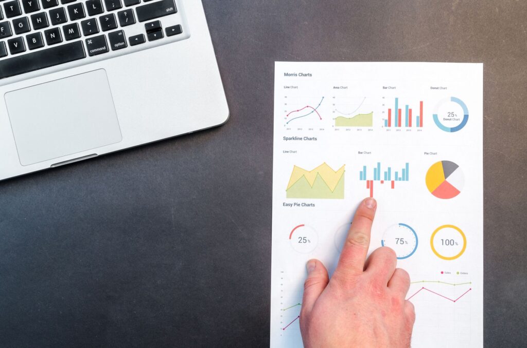 A hand pointing at colorful charts with a laptop in the background on a desk.