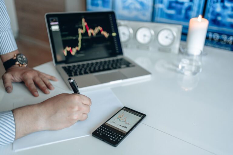Business analyst writing with laptop and smartphone showing trend graphs on a white desk.