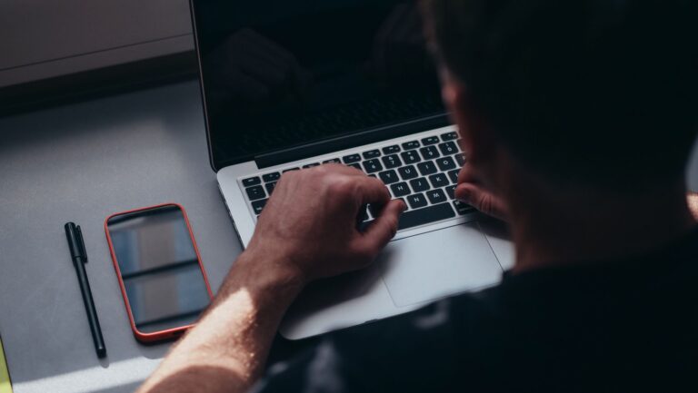 Close-up of a man working on a laptop with a smartphone and pen on the table.